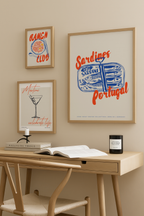 Ett litet träbord med en stol står mot en beige vägg dekorerad med Bozettos Bundle 3-pack Kökstavlor No. 1, med inramade bilder på sardiner, ramen och en martini. Ett ljus och en bok vilar på skrivbordet.