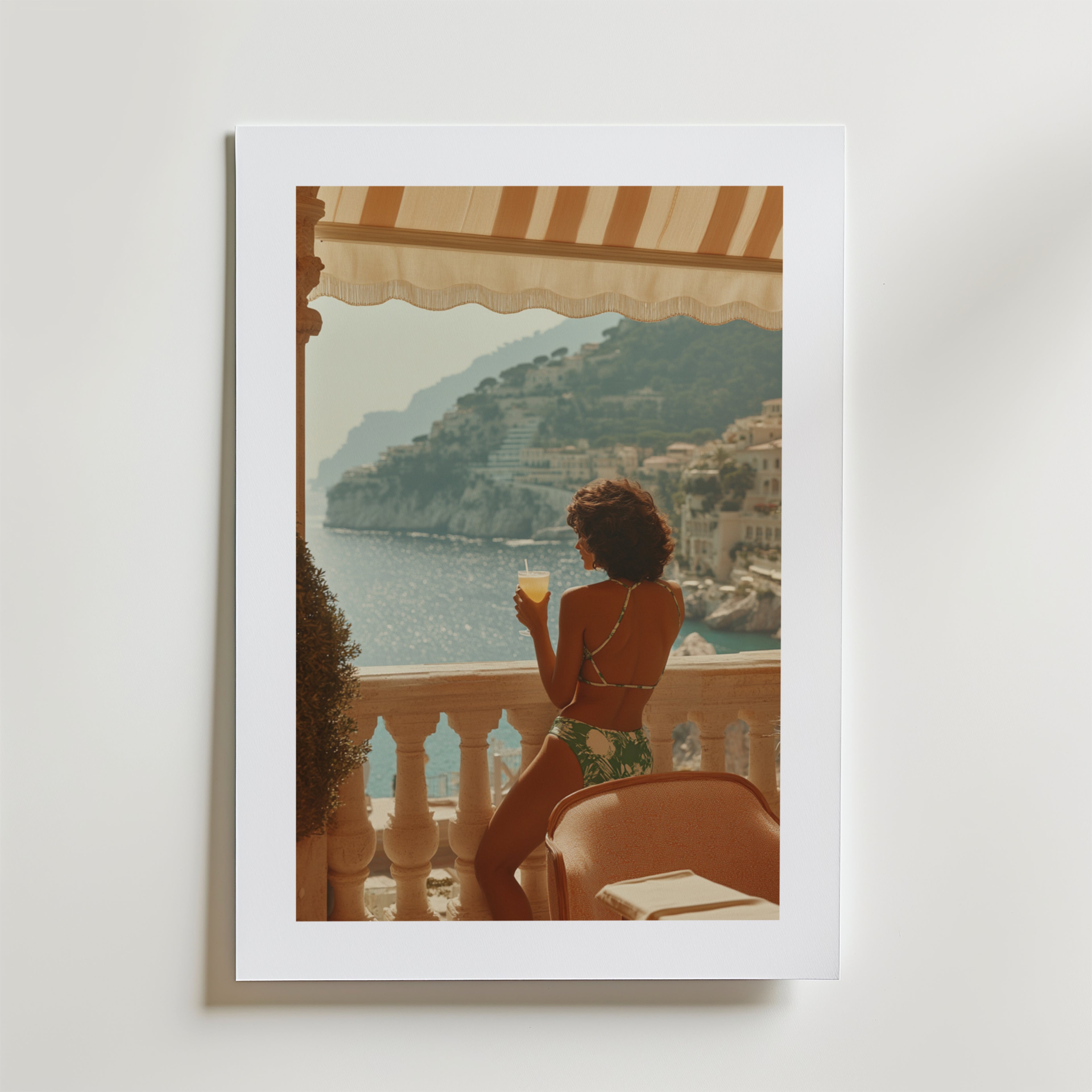 En kvinna i baddräkt står på en balkong med utsikt över havet och klipporna och håller i en drink under en randig markis. Medelhavsbyggnader pryder kullarna på denna högkvalitativa Amalfi Coast Balcony Poster by Bozetto.