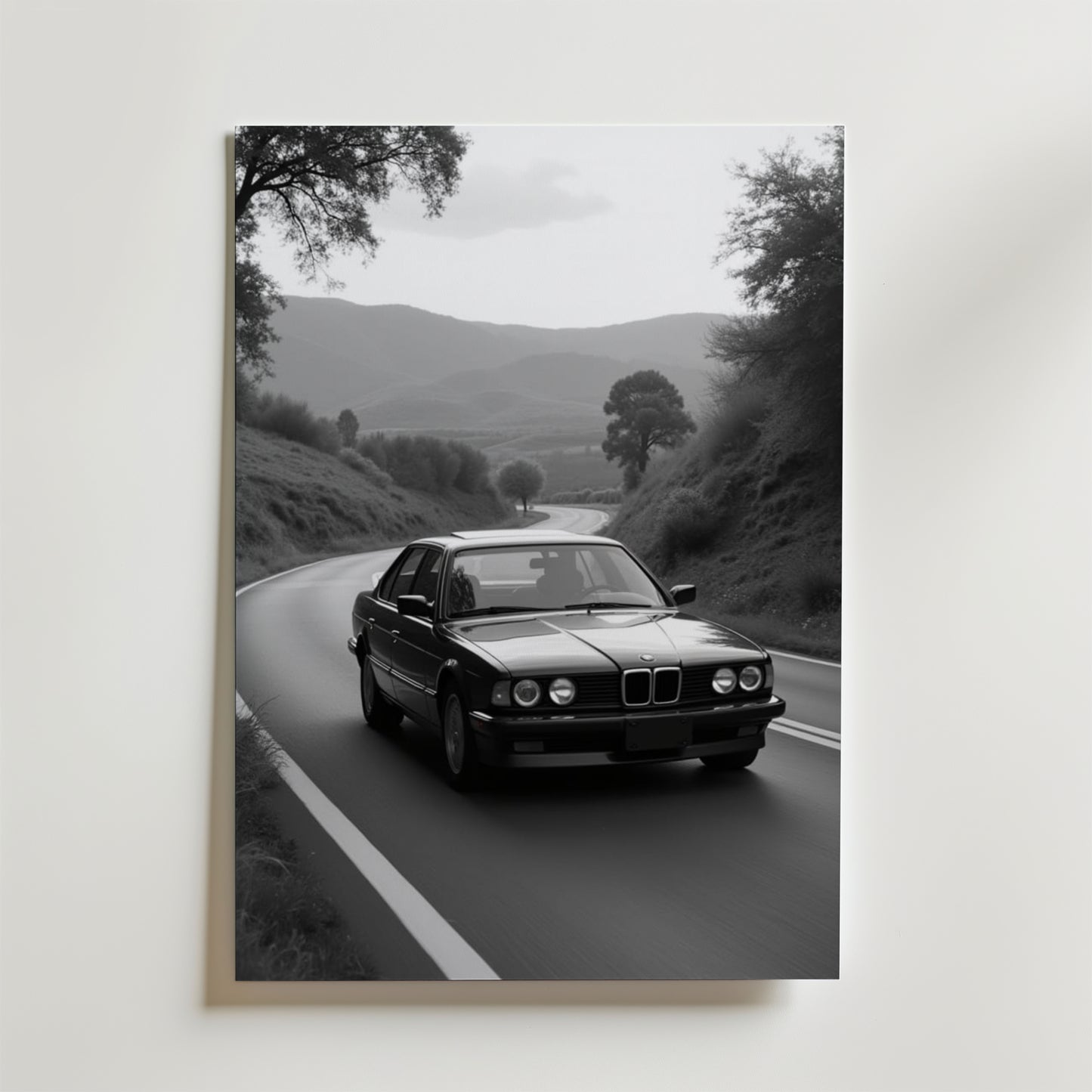 Vintage BMW Poster by Bozetto: Svartvitt foto av en klassisk sedan som kör på en slingrande väg genom kullar, omgiven av träd och berg, tryckt på premiumpapper med en lyxig matt finish.