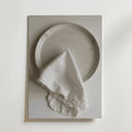 En Bozetto White linen dinner napkin Poster visar en vit keramiktallrik som toppas med en fransig vit linneservett, allt mot en vit bakgrund med en lyxig matt finish.
